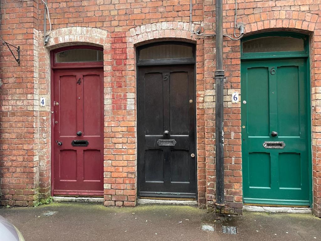 Three doors in a row on a brick wall; left door is maroon, middle door is black, and right door is green. Each door has a number on its upper left side.